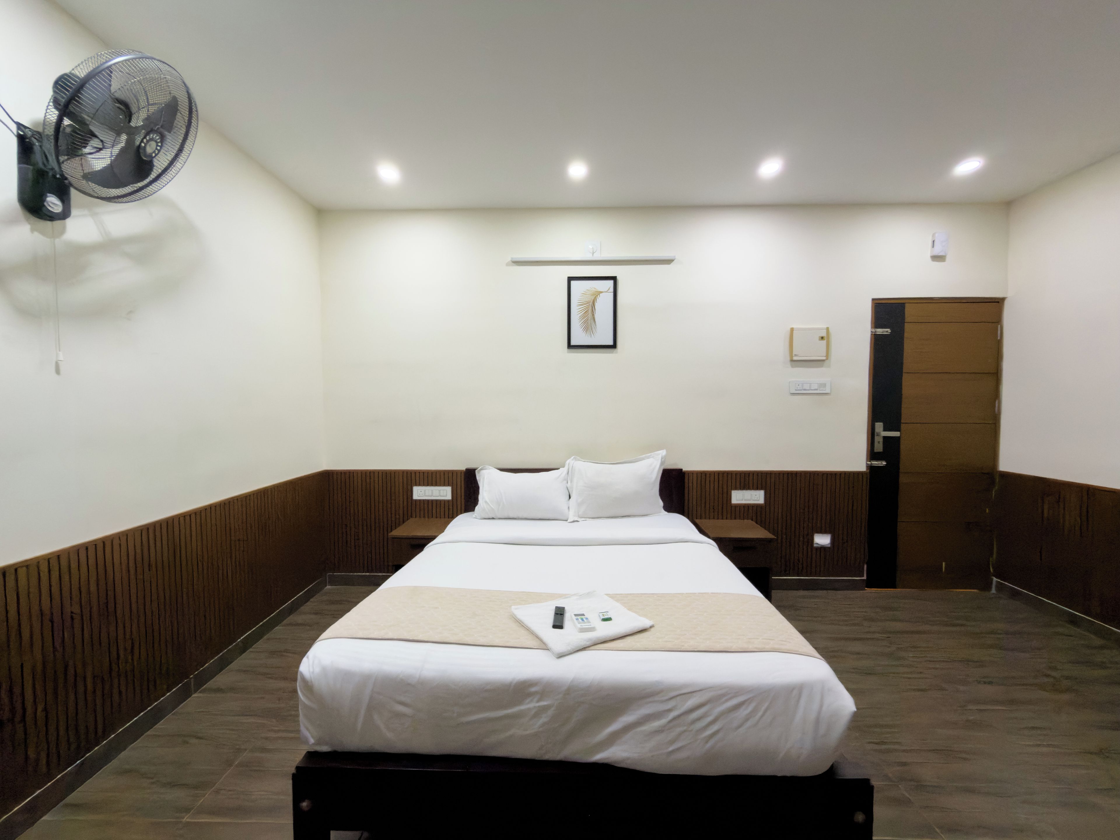 A hotel room with a bed, bedside tables, a wall-mounted fan, a door, and a framed picture on the wall.