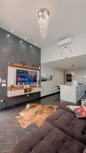 A modern living room with a large flat-screen TV, a leather sofa, a cowhide rug, and a chandelier. - Foz do Iguaçu House for the Comfort of your Family (Residencial Cohigaçu Foz do Iguaçu)