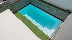 Una piscina rectangular con agua azul cristalina, rodeada por un borde de césped artificial verde y un muro de piedra.
