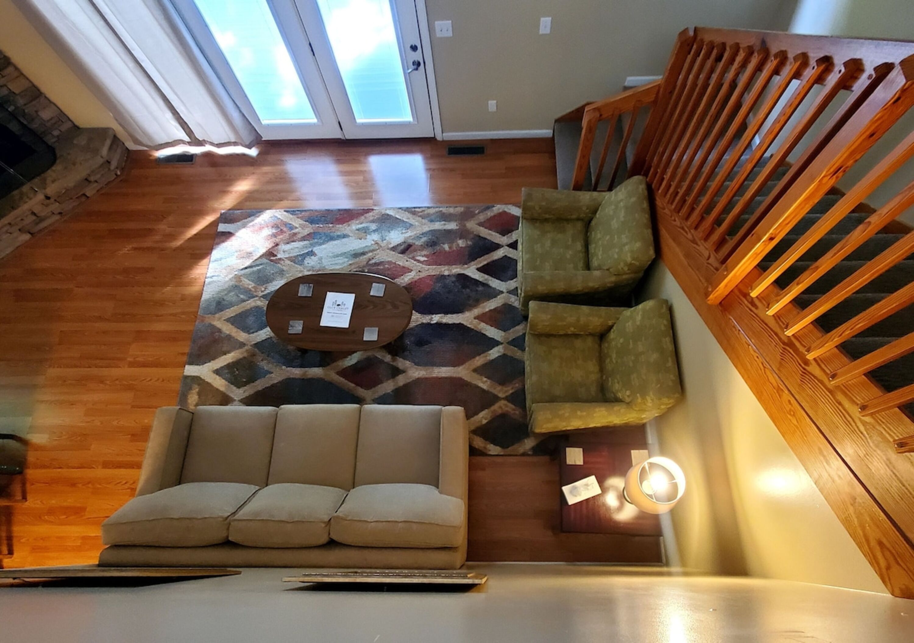 Ein Wohnzimmer mit einer Couch, zwei Sesseln, einem Couchtisch und einem gemusterten Teppich. Im Hintergrund ist eine Holztreppe zu sehen.