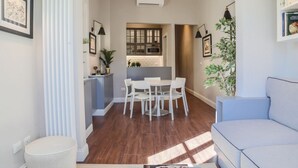 Un salon moderne avec un canapé gris, une table à manger avec des chaises blanches, un petit pouf rond et un coin cuisine visible à travers une porte ouverte.