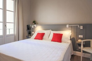 Una cama bien tendida con almohadas blancas y rojas, una mesita de noche con una lámpara y una planta pequeña en un estante.