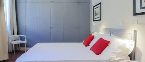 A neatly made bed with white and red pillows, a white chair, and a small table with a book.