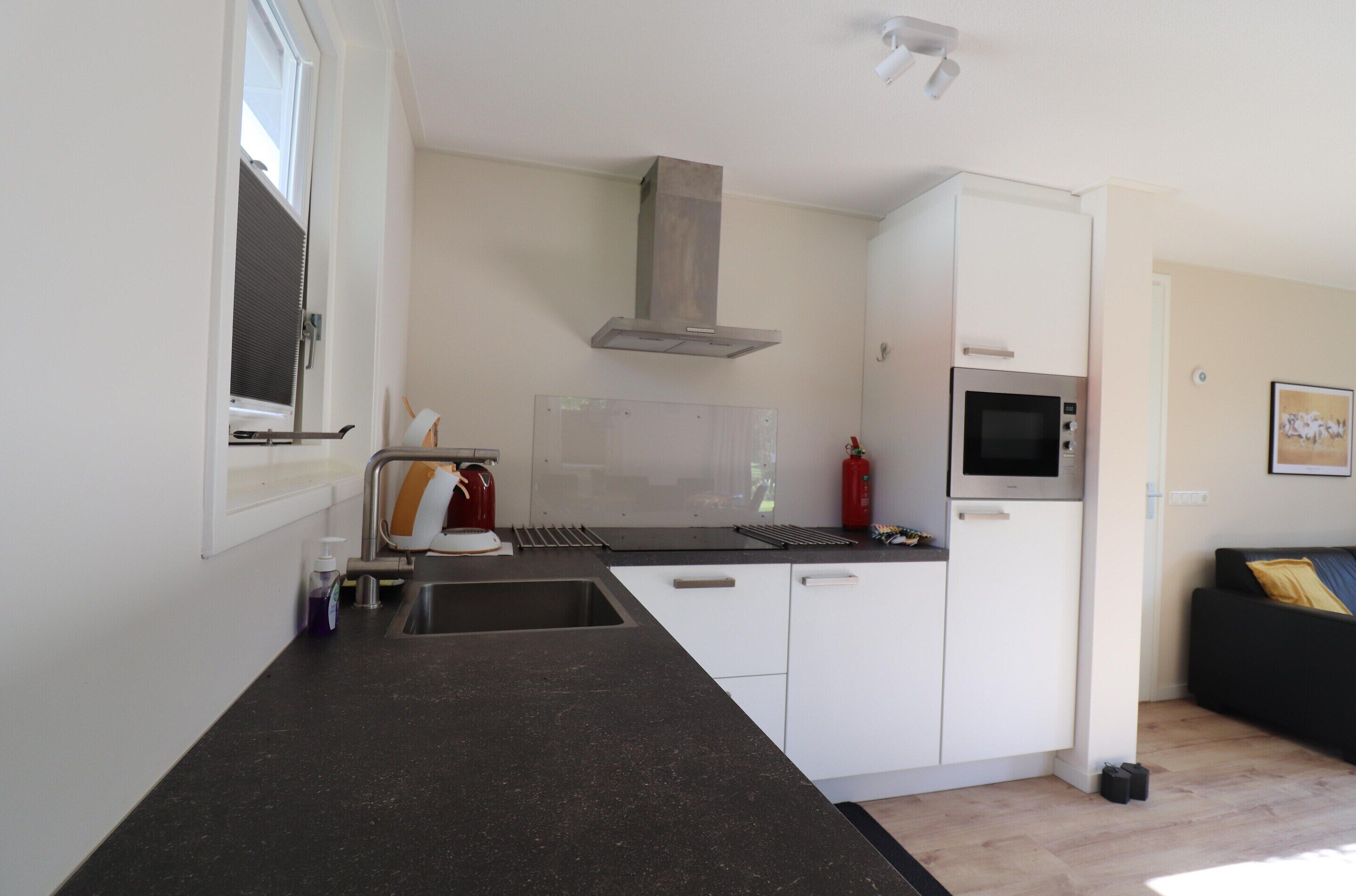 A compact kitchen with a microwave, stove, and sink. There is a window with blinds, a fire extinguisher, and a black countertop.