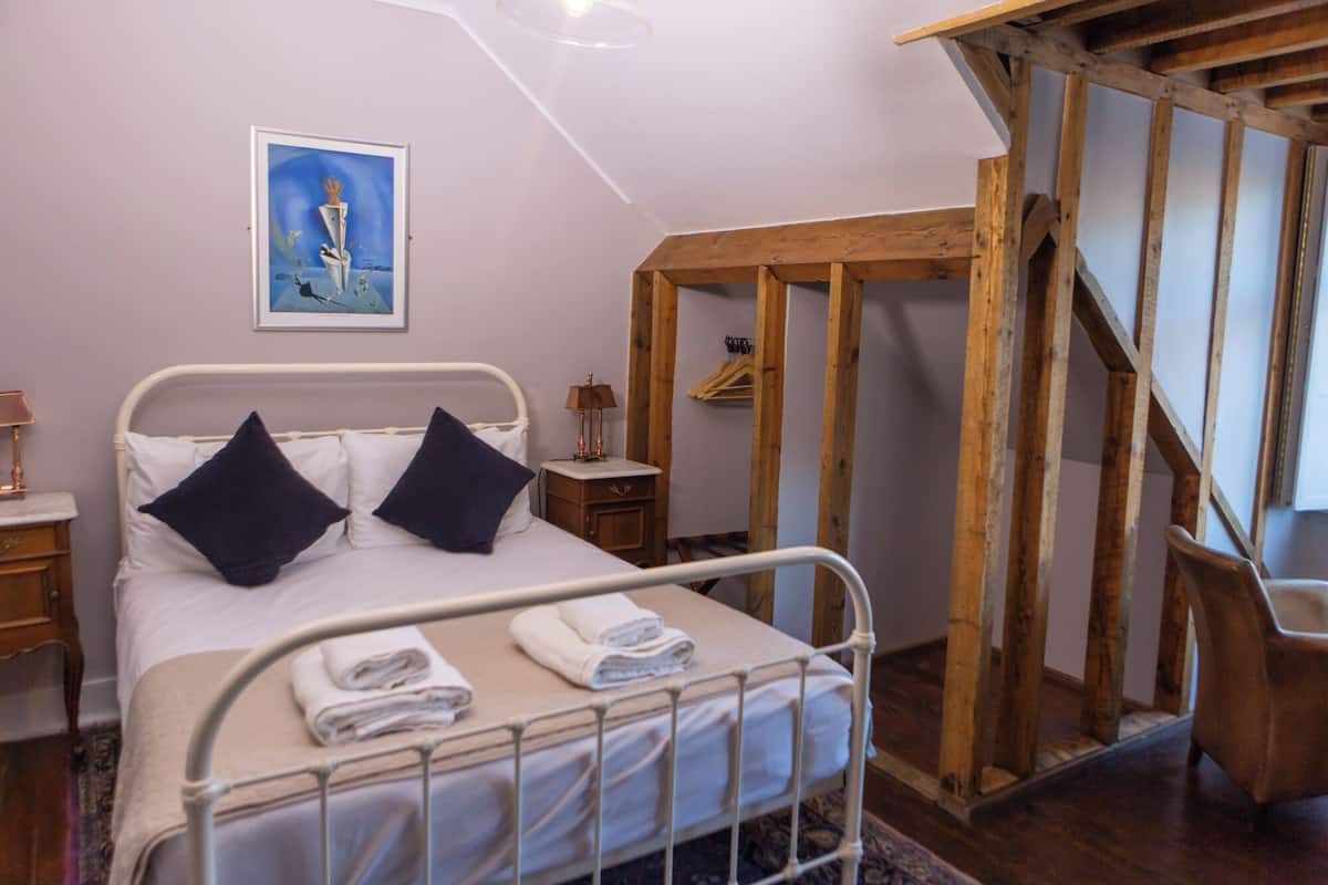 A bedroom with a metal bed frame, two pillows, a bedside table with a lamp, a wooden staircase, and a framed picture on the wall.