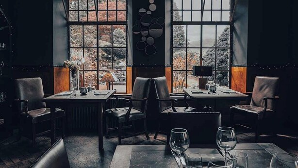 A cozy restaurant with two tables and chairs, large windows with a view of trees, and a wall-mounted circular decoration. - Haweswater Hotel (Penrith)