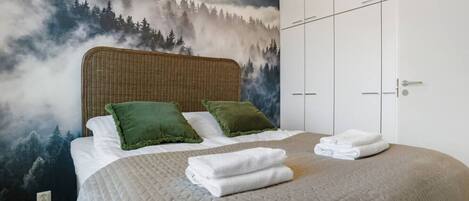 Une chambre à coucher avec un lit, des oreillers verts, une couvre-lit matelassée et une fresque représentant une forêt sur le mur.