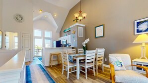 A spacious kitchen with white cabinets, a dining area with a wooden table and chairs, and a living area with a sofa and a lamp. - 2 BR Condo with shared pool and central air (Kill Devil Hills)
