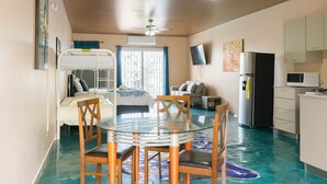 Ein Zimmer mit Etagenbett, einem Glastisch mit Stühlen, einem Deckenventilator, einer Küchenzeile inklusive Kühlschrank und Mikrowelle sowie einem großen Fenster mit Vorhängen.