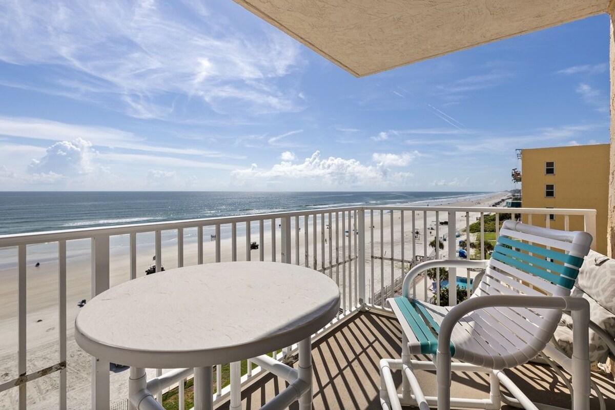 Un balcon avec une table blanche et deux chaises, donnant sur une plage sous un ciel dégagé.