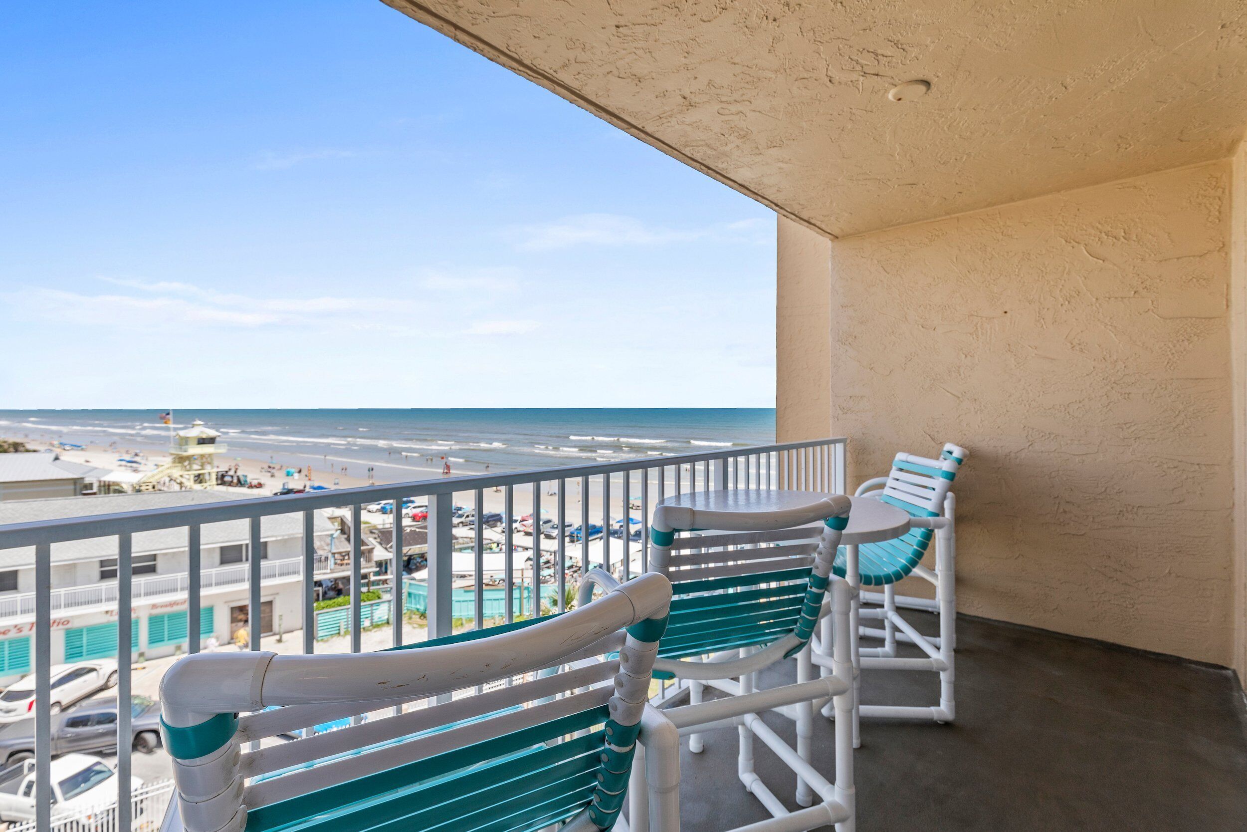 A balcony with two white chairs and a table, overlooking a beach and a parking area.
