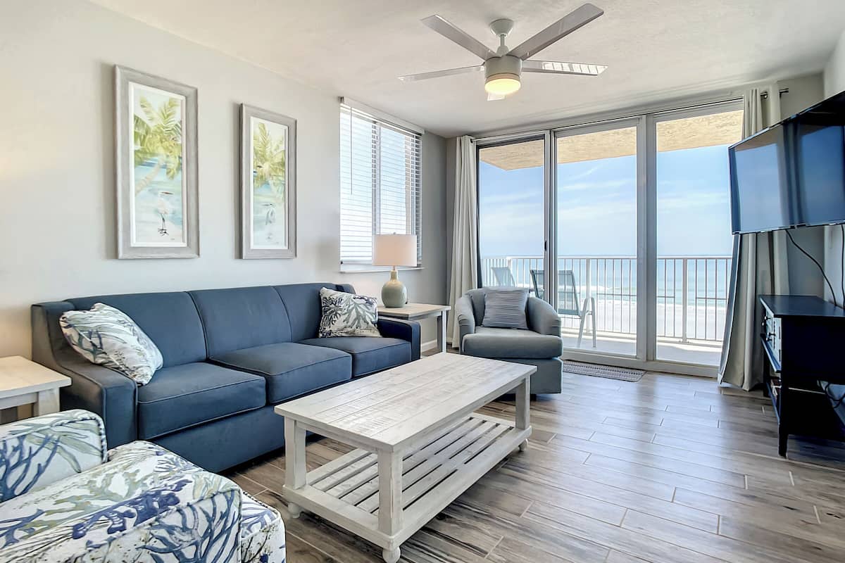 Un vaste espace de vie comprenant un canapé bleu, une table basse blanche et un fauteuil gris. Deux tableaux encadrés ornent le mur, ainsi qu’un ventilateur de plafond et une grande fenêtre offrant une vue sur la plage.