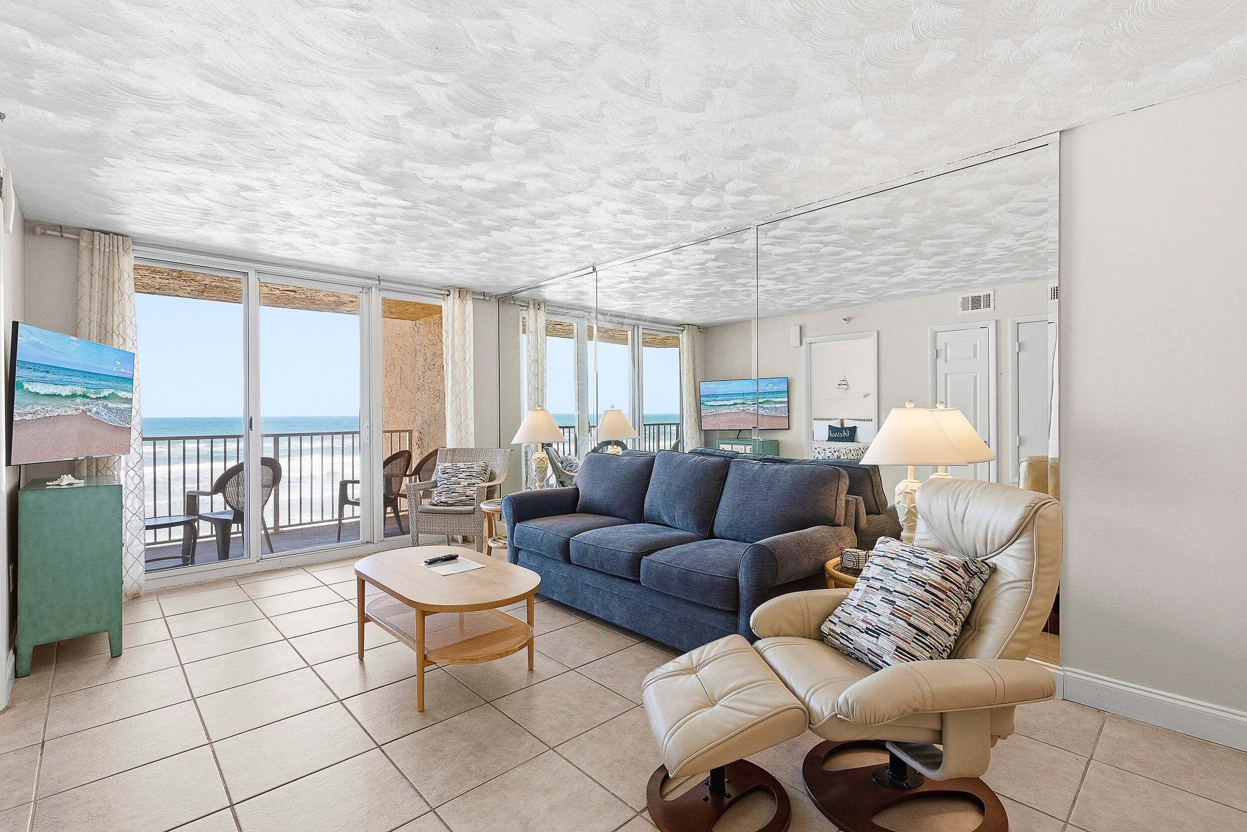 A living room with a sofa, armchair, and a table, a balcony with a view of the sea, and a ceiling with a textured pattern.