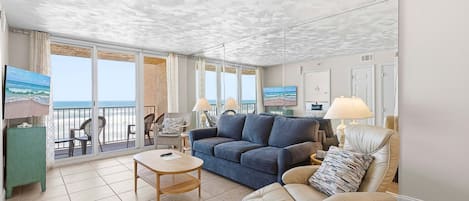 Ein Wohnzimmer mit einer Couch, einem Sessel und einem Tisch, ein Balkon mit Meerblick und eine Decke mit strukturierter Musterung.