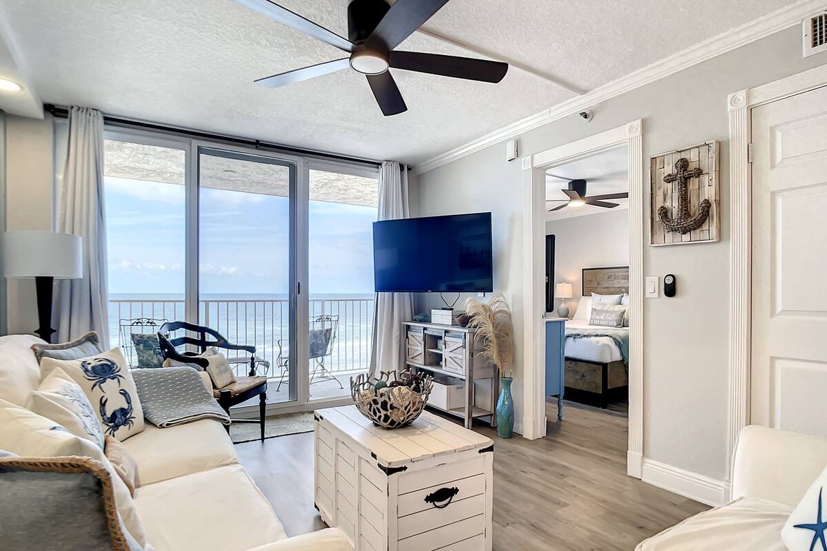 Ein Wohnzimmer mit einem weißen Sofa, einem Fernseher, einem Deckenventilator und Meerblick durch große Fenster.