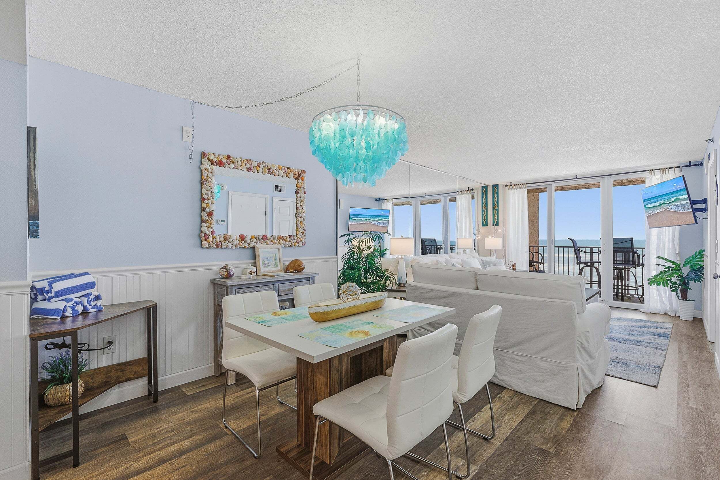 A modern living room with a dining area, a large chandelier, a white sofa, and a view of the ocean through large windows.