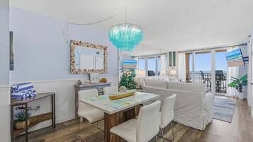 A modern living room with a dining area, a large chandelier, a white sofa, and a view of the ocean through large windows.