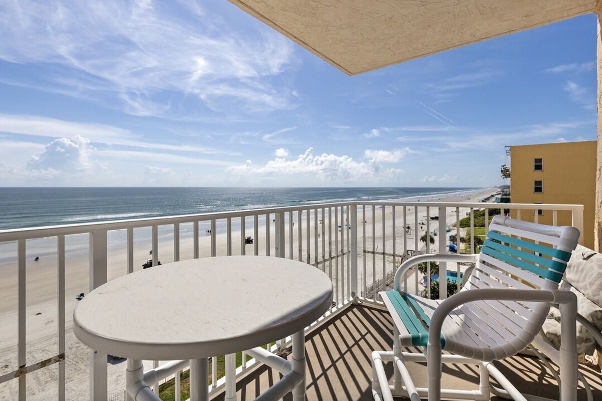 Ein Balkon mit einem weißen Tisch und zwei Stühlen mit Blick auf einen Strand unter blauem Himmel.