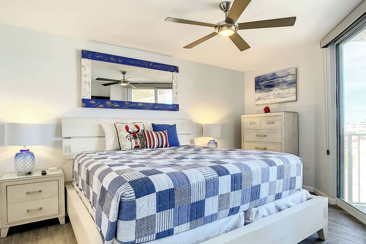 Une chambre avec un grand lit, un ventilateur de plafond, une table de chevet avec une lampe, une commode et une fenêtre donnant sur un beau paysage.