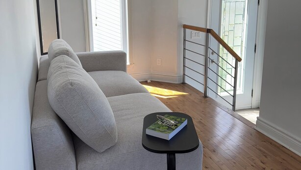 A living room with a grey sofa, a small round table with a book, a staircase, and a door leading outside.