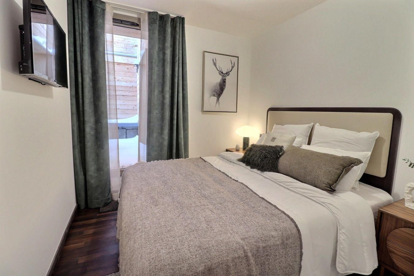 Ein Schlafzimmer mit einem Bett, einem an der Wand montierten Fernseher, einem Fenster mit Vorhängen und einem Bild von einem Reh an der Wand.