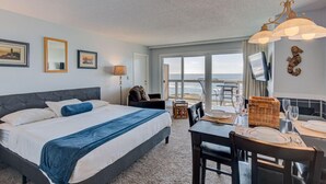 A hotel room with a bed, a dining table set for two, a TV, and a view of the ocean. - Panoramic Ocean View Studio with Full Kitchen (Otter Rock)