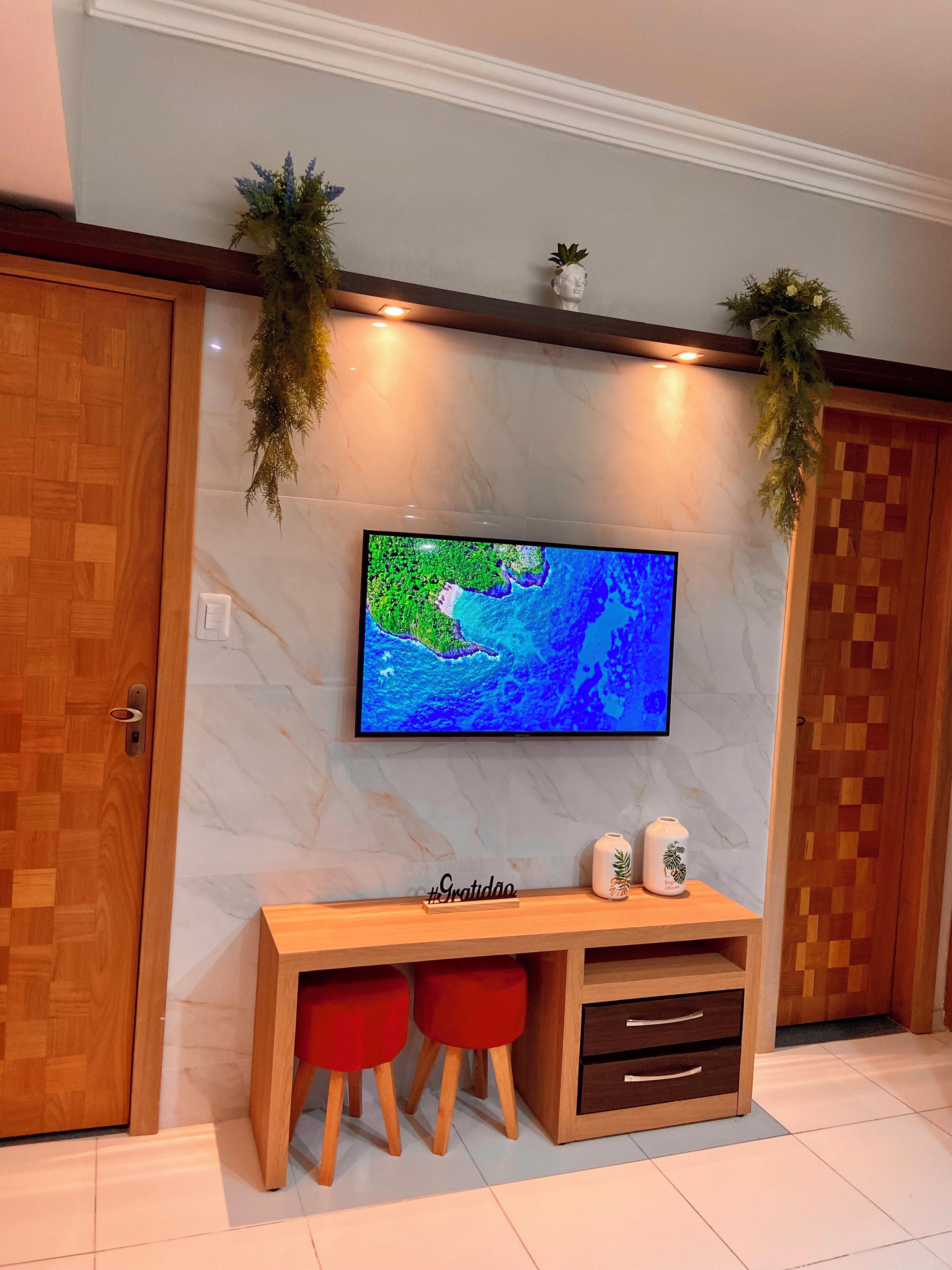 A room with a television mounted on a marble wall, a wooden desk with two red stools, and a cabinet with drawers.
