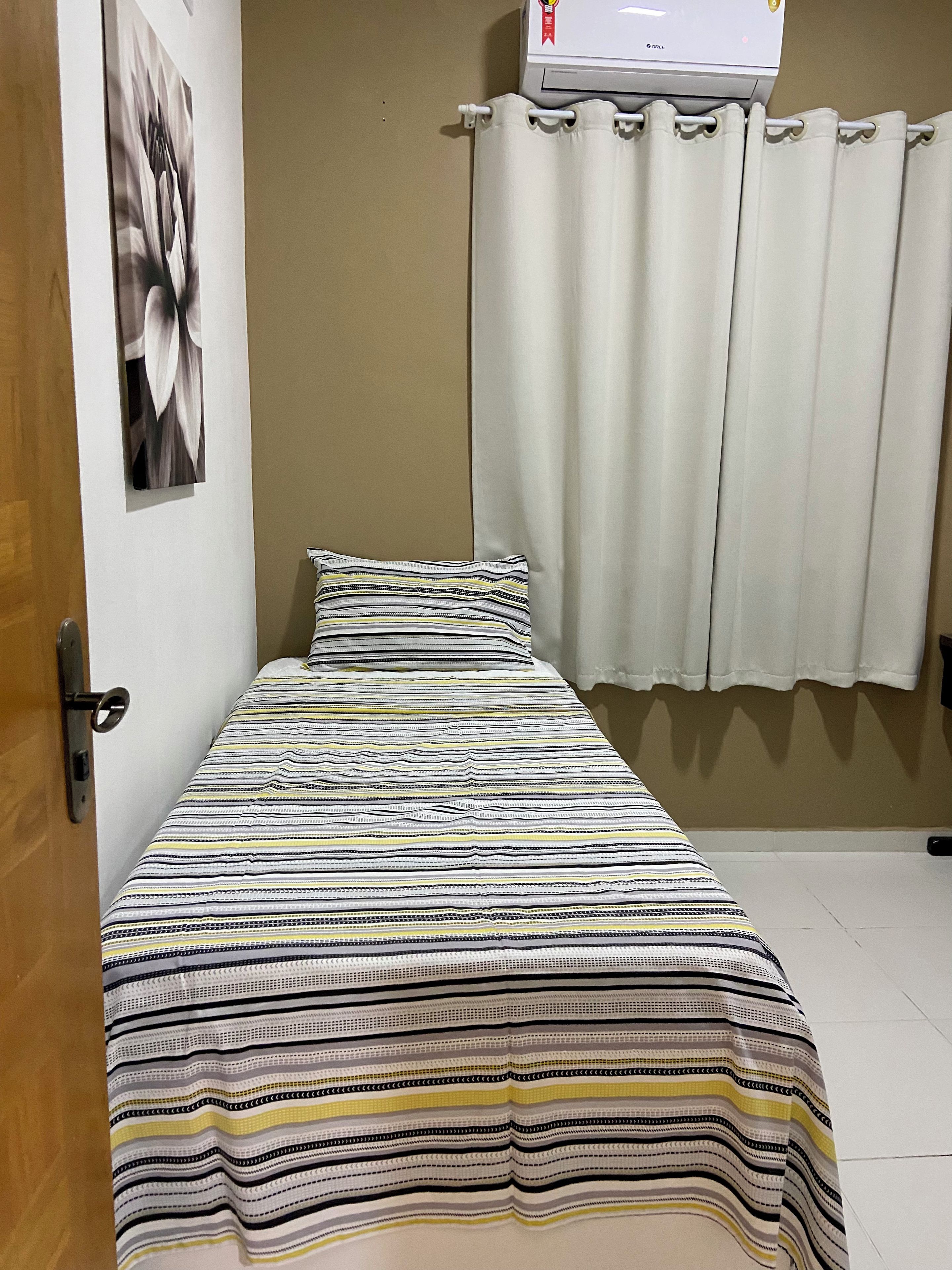 Una cama individual con un edredón a rayas, una almohada, un aire acondicionado de pared y una ventana con cortinas blancas.
