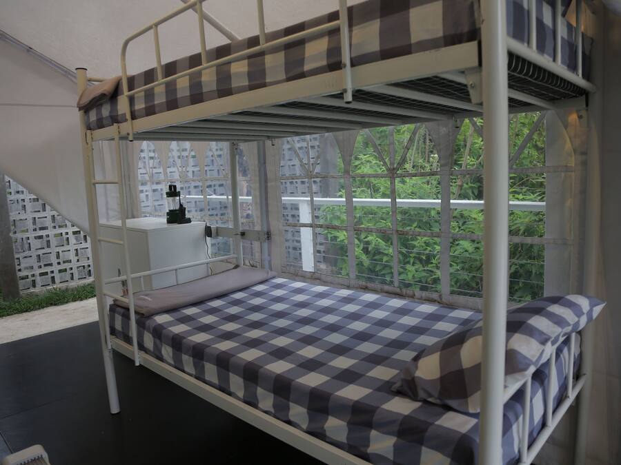A bunk bed with a checkered mattress, a small desk with a chair, and a window with a view of greenery and buildings.