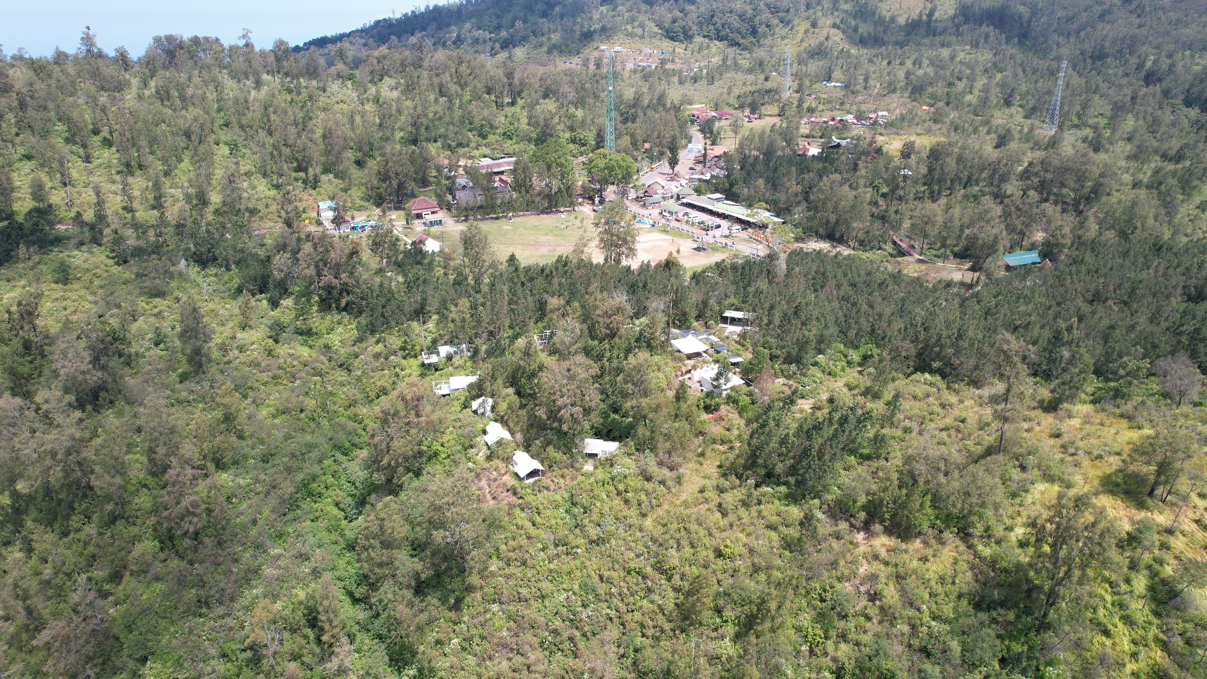 Eine Vogelperspektive zeigt eine kleine Stadt, die von dichtem Wald umgeben ist, mit mehreren Gebäuden und einer großen Struktur, die vermutlich ein Gemeinschaftszentrum oder eine ähnliche Einrichtung ist.