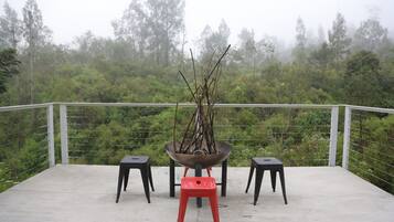 A balcony with a concrete floor, two black stools, a red stool, and a metal fire pit with branches.