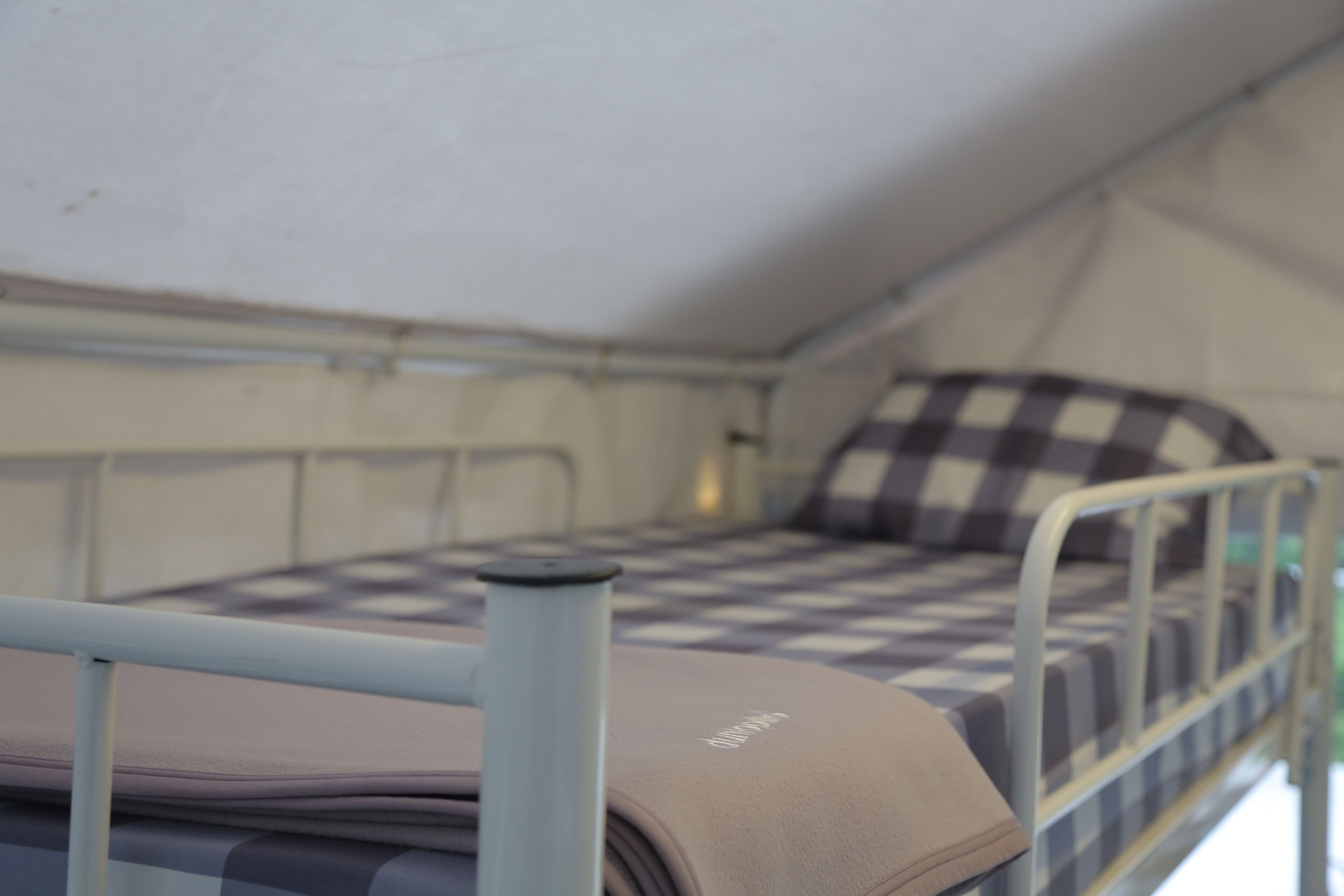 A bunk bed with a checkered mattress and a pillow, a metal railing, and a tent-like structure in the background.