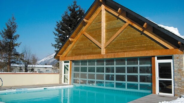 Un edificio de madera con puerta de garaje, piscina y montaña al fondo.