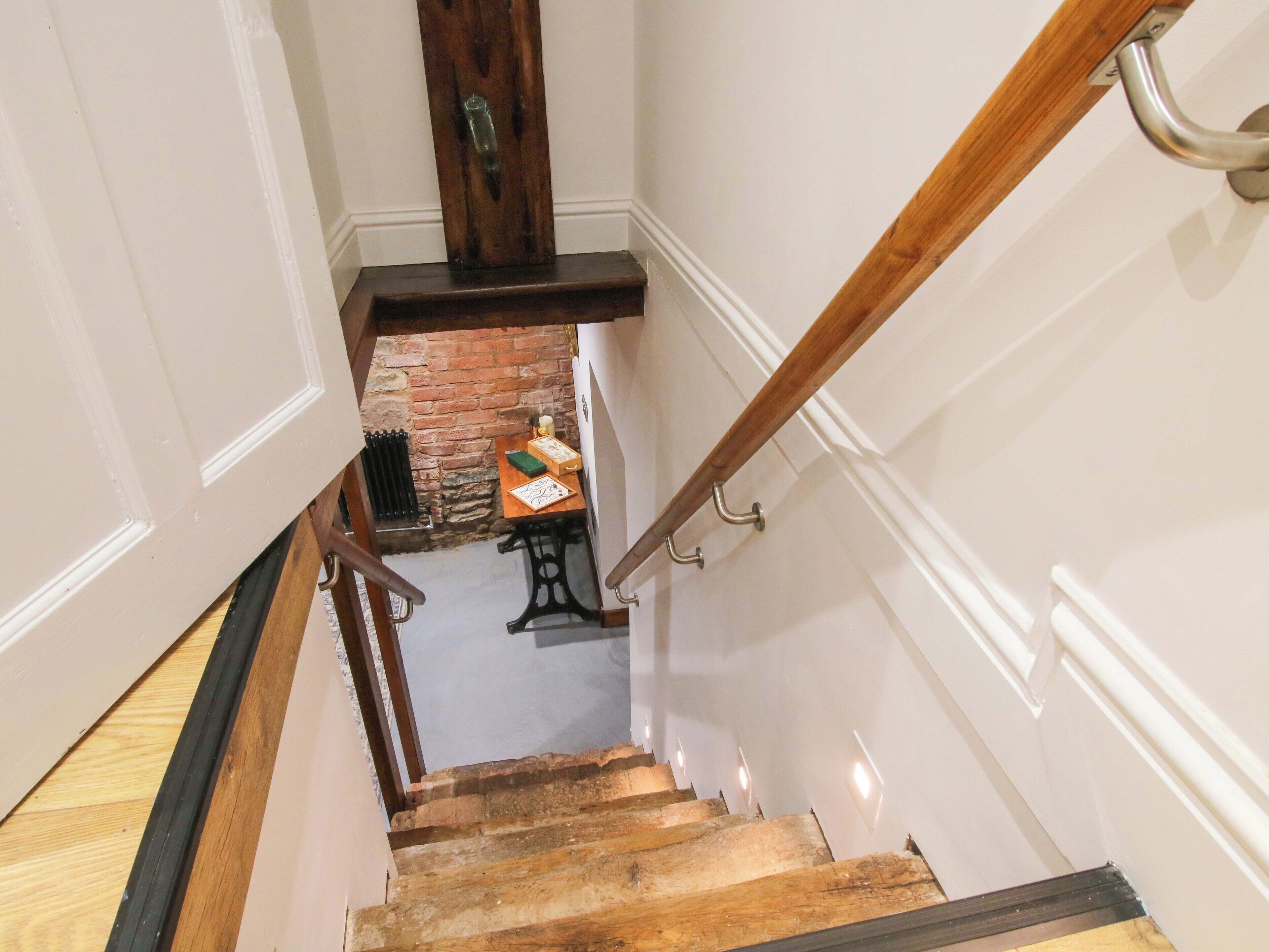 A wooden staircase with a handrail, leading to a room with a brick wall and a table with a lamp.