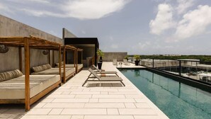 A rooftop pool area with a concrete wall, a wooden pergola with seating, and a clear sky.