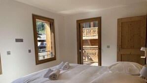 A bedroom with a large bed, two windows, a wooden door, and a balcony with a railing. - 100m from resort center, 2nd floor, view mountain/village, balcony, tv, 69m², Morzine (Morzine)