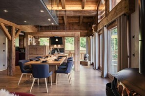 A spacious dining area with a long wooden table and blue chairs, a kitchen with a large island, and large windows offering a view of the outdoors. - Chalet, view panoramic/mountain, terrace, balcony, fireplace or stove, parking, tv, 317m², La Clusaz (La Clusaz)
