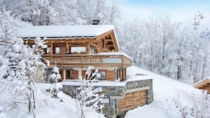 Une cabane en bois, posée sur des fondations en pierre, entourée d’arbres couverts de neige et avec une chaîne de montagnes en toile de fond.