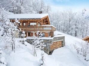 Eine Holzhütte mit steinernen Fundamenten, umgeben von schneebedeckten Bäumen und einer Bergkulisse.