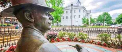 A statue of a man in a hat pointing to the right, surrounded by a colorful garden and a white building in the background.