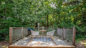 A wooden deck with a round table and two chairs, surrounded by a wooden railing, set in a wooded area. - The Pappy, 2BR House by Velvet Ditch Villas (Oxford)