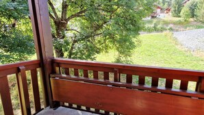 Une terrasse en bois avec un banc, donnant sur une zone herbeuse bordée d’arbres et sur laquelle on aperçoit un bâtiment au loin.