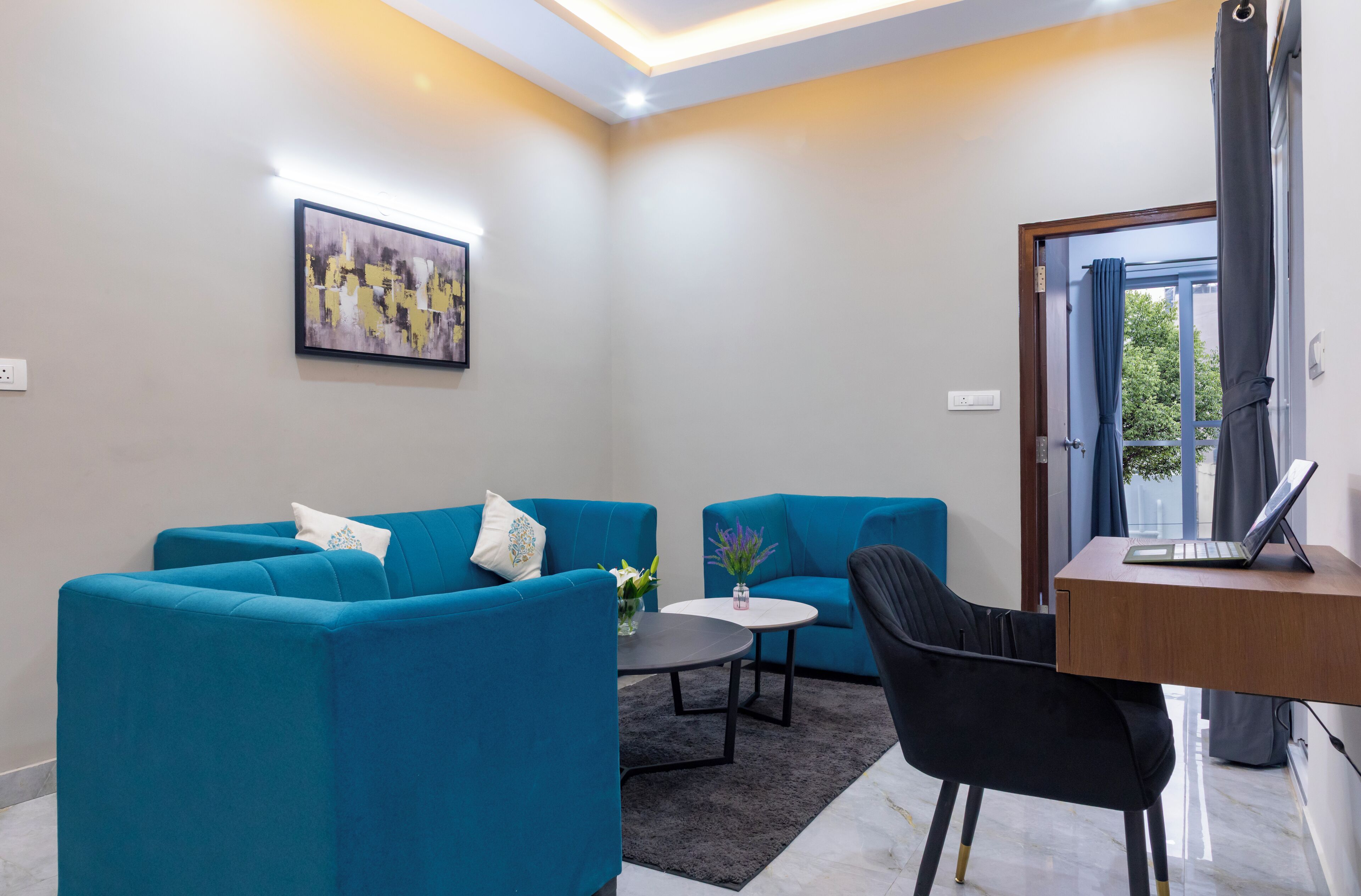 A modern living room with two blue sofas, a small round table, a black chair, and a laptop on a wooden desk.