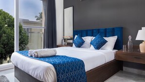 Un dormitorio moderno con una cama grande, cabecera azul oscuro y vistas a palmeras a través de una ventana.