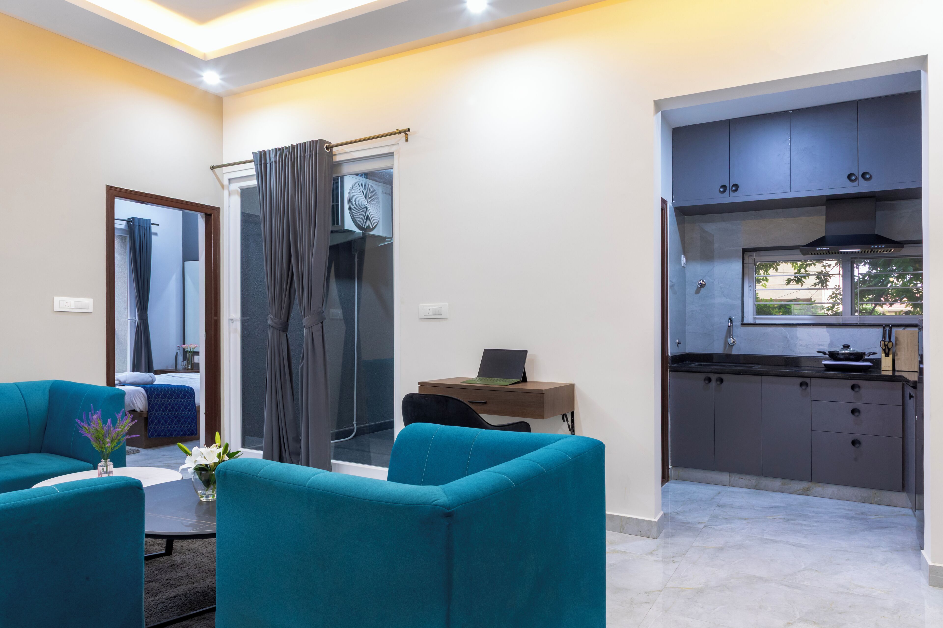A modern living room with teal sofas, a coffee table, and a laptop on a wooden side table. A glass door leads to a shower area with a towel rack.
