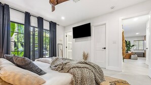 A bedroom with a large bed, a TV mounted on the wall, and a view of the outdoors through the window. - Luxury Waterfront 2BR in Wilton Manors • Jungle-Luxe Retreat • TABŪ (Fort Lauderdale)
