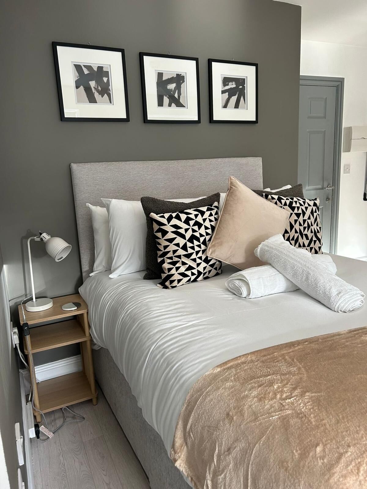 A neatly made bed with a grey headboard, a bedside table with a lamp, and three framed artworks on the wall.