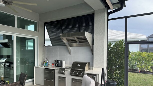 Et udendørs køkken med grill, vask og skabe, beliggende ved siden af en glasdør, der fører til en terrasse med fletmøbler.