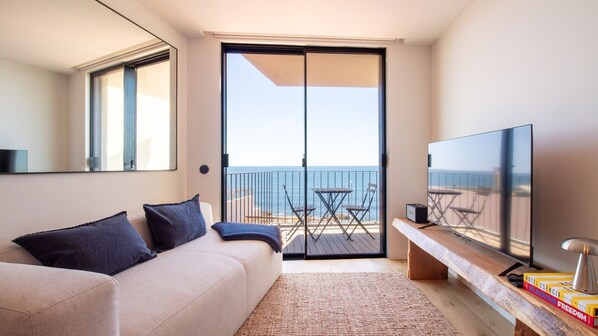 Una camera d'albergo moderna con divano, TV a schermo piatto, una panca in legno e un balcone con vista sul mare.