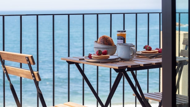 Una mesa de madera con alimentos para el desayuno, un bol de fresas, una taza y un tarro de mermelada, con vistas al mar.